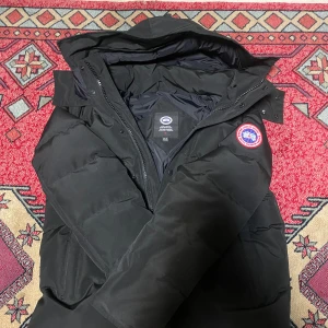 Svart dunjacka från Canada Goose - Säljer en svart dunjacka i storlek M. Jackan är perfekt för kalla vinterdagar med sitt varma dunfoder. Den har en fin design med en huva och en tydlig logotyp på ärmen. Jackan är i nyskick och redo för nya äventyr i kylan!