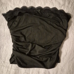 Svart ribbad topp med spetsdetaljer - Säljer en snygg svart ribbad tubtopp med vacker spetskant och en liten rosett framtill. Perfekt för en kväll ute eller en festlig tillställning. Toppen är axelbandslös och har en tight passform som framhäver figuren.