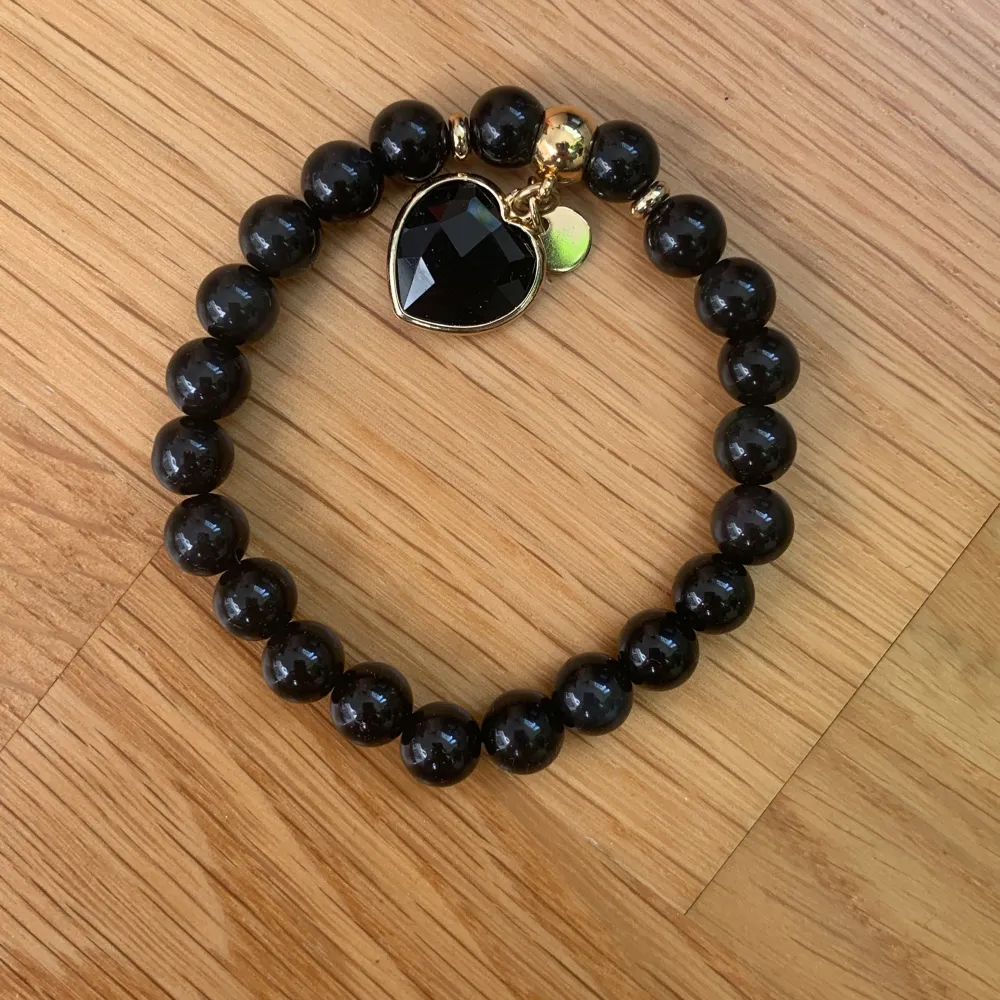 Snyggt svart pärlarmband i obsidian med ett stort hjärthänge och en liten guldfärgad detalj. Perfekt för att ge en elegant touch till din outfit. Armbandet är elastiskt och passar de flesta handleder.. Accessoarer.