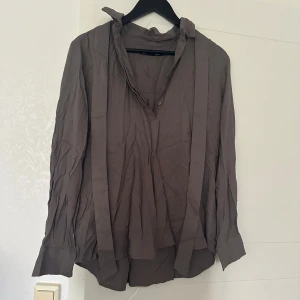 Skjorta/blus - Skjorta/blus med knytning. Bandet är avtagbart (se bild). Svårt att få rättvis bild på färgen men där är grå/grön. Endast använd ett fåtal gånger så den är hel och fräsch!