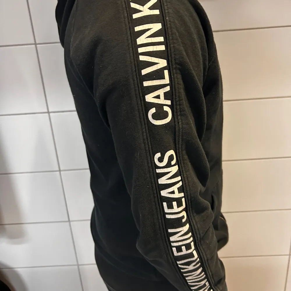 Säljer en snygg svart hoodie från Calvin Klein Jeans. Den har en dragkedja framtill och text längs ärmarna. Perfekt för en casual look eller när du bara vill vara bekväm. Den är i bra skick och redo för en ny ägare!. Tröjor & Koftor.