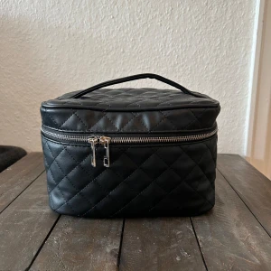 Svart quiltad necessär - Säljer en snygg och praktisk svart quiltad necessär. Den har en dragkedja som går runt hela väskan och ett handtag upptill för enkel transport. Perfekt för att organisera dina toalettartiklar eller smink när du är på resande fot!