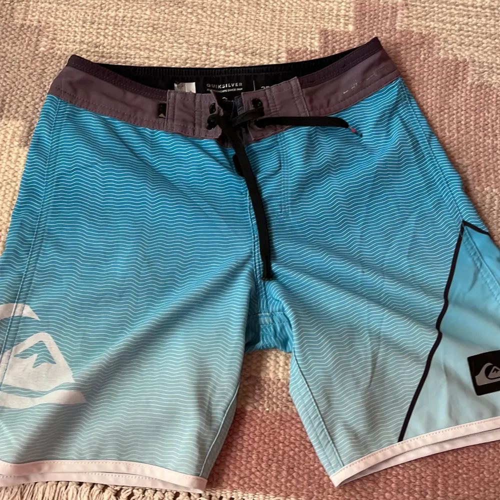 Säljer ett par snygga blå boardshorts från Quiksilver. De har ett coolt vågmönster och en svart dragsko i midjan. Perfekta för sommarens alla strandäventyr! 🏄‍♂️. Shorts.