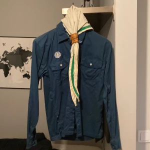 Blå scoutskjorta med halsduk - Säljer en blå scoutskjorta med en vit och grön halsduk. Skjortan har långa ärmar och två bröstfickor med knappar. Perfekt för scoutäventyr eller som en cool vintage-stil. Storlek 164/170, 