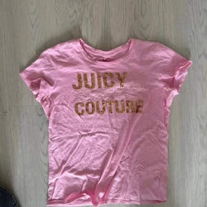 Rosa t-shirt från Juicy Couture - Säljer en superfin rosa t-shirt från Juicy Couture. Den har korta ärmar och ett glittrigt guldtryck med märkesnamnet på framsidan. Perfekt för en avslappnad men ändå stilren look. T-shirten är i bomull och känns riktigt skön mot huden.