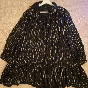 Svart klänning med guldmönster - Snygg svart klänning med guldmönster och volangdetaljer. Klänningen har långa ärmar och knappar framtill. Använd på ett sparsamt sett