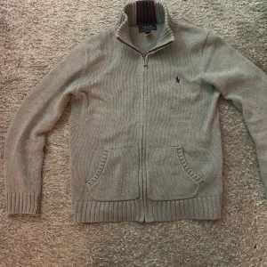Grå kofta från Polo Ralph Lauren - Snygg grå kofta från Polo Ralph Lauren med dragkedja och ribbad krage. Den har två praktiska fickor framtill och ett diskret broderat märke på bröstet. Perfekt för kyligare dagar! Storleken M i barn.