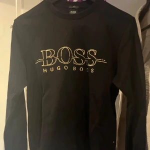 Hugo Boss Crewneck - Snygg svart långärmad tröja från Hugo Boss med guldtryck på bröstet. Perfekt för en stilren och avslappnad look. Tröjan har en klassisk passform och rund halsringning. Mycket bra skick!
