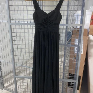 Svart långklänning med rynkdetaljer - Elegant svart långklänning med breda axelband och vacker rynkdetalj vid bysten. Klänningen har en flytande passform och är perfekt för festliga tillfällen. Materialet ger en fin glans och faller snyggt.