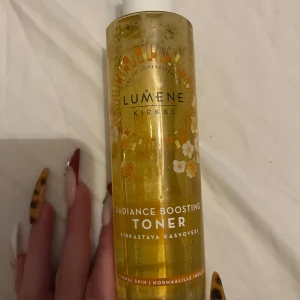 Lumene Kirkas Radiance Boosting Toner - Fräsch och uppfriskande toner från Lumene Kirkas som ger huden en strålande lyster. Perfekt för normal hud och innehåller naturliga ingredienser för en skonsam rengöring. Flaskan är genomskinlig med en vit kork och har en gyllene etikett med blommönster.