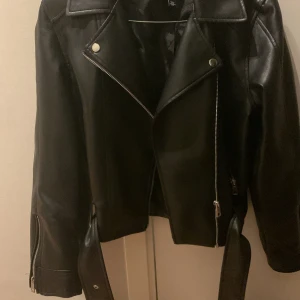 Svart skinnjacka med dragkedja - Snygg svart skinnjacka med dragkedja och knappar. Jackan har en klassisk bikerstil med långa ärmar och detaljer som ger en cool look. Perfekt för att ge din outfit en edgy touch.