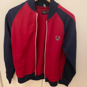 Röd och marinblå kofta från Fred Perry - Snygg kofta i rött och marinblått från Fred Perry. Koftan har en dragkedja framtill och ribbade muddar vid ärmslut och nederkant. Den ikoniska Fred Perry-loggan pryder bröstet.