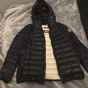 Svart dunjacka från Parajumpers - Snygg svart dunjacka från Parajumpers med huva och dragkedja. Jackan har ett emblem på ärmen och quiltad design. Perfekt för kyliga dagar.