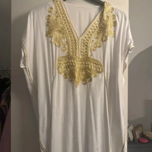 Vit kaftan med guldbroderi - Elegant vit kaftan med detaljerat guldbroderi runt halsen. Den har en lös passform och korta ärmar, perfekt för en stilfull look. Broderiet ger en lyxig känsla och gör plagget unikt.