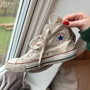 Vita Converse All Star sneakers - Säljer ett par klassiska vita Converse All Star sneakers med snörning. Skorna har en hög modell med det ikoniska märket på sidan och en röd och blå rand runt sulan. I användt bruk. Stl 6. Stl 39. 