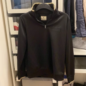 Svart tröja från Illusive London - Snygg svart tröja från Illusive London med dragkedja vid halsen. Perfekt för en stilren look. Tröjan har långa ärmar och en diskret logga på bröstet. Produkten är i bra skick och är i storlek XS