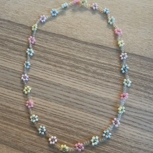 Färgglatt blommigt halsband - Charmigt halsband med små blommor i pastellfärger som rosa, blå, gul och lila. Perfekt för att ge en lekfull touch till din outfit. Blommorna är jämnt fördelade längs hela halsbandet och ger en söt och somrig känsla.