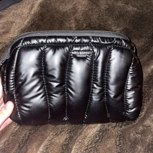 Svart puffer necessär/väska från ART STREET LONDON - Snygg svart puffig Necessär/väska från ART STREET LONDON med dragkedja. Perfekt för att ha dina saker när du reser kort eller annat. Den har en mjuk och glansig yta som ger en modern touch. Har 3st innefack, en av dem har dragkedja. Köpt på ullared. 