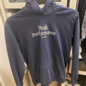 Mörkblå hoodie från Peak Performance - Säljer en snygg mörkblå hoodie från Peak Performance med vit text på bröstet. Tröjan har en klassisk känguruficka och en bekväm huva. Perfekt för en avslappnad stil. Hoodien är i bra skick och är i storlek 170