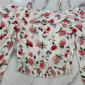 Blommig offshoulder topp från H&M - Säljer en söt blommig offshoulder topp från H&M. Toppen har ett färgglatt blommönster i rött, rosa och lila. Den har långa ärmar och en volangdetalj vid axlarna. Perfekt för vår och sommar!
