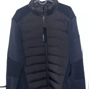 Svart jacka från Sail Racing - Snygg svart jacka från Sail Racing med dragkedja framtill. Jackan har en pufferdesign på framsidan och är tillverkad i ett syntetmaterial. Perfekt för kyligare dagar med sin stilrena look och bekväma passform.