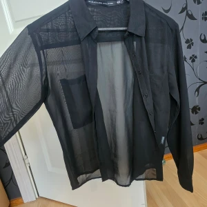 Svart transparent skjorta från Black Label Collection - Snygg svart transparent skjorta från Black Label Collection. Skjortan har långa ärmar och knappar framtill. Perfekt för en stilren och modern look.
