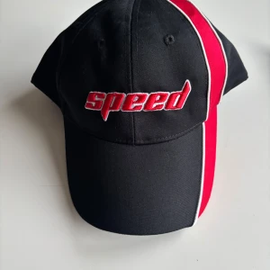 Svart och röd keps med texten 'Speed' - Snygg svart keps med röd och vit rand och texten 'Speed' broderad framtill. Perfekt för en sportig look. Justerbar passform för optimal komfort.