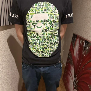 Svart t-shirt från A Bathing Ape - PRIS GÅR ATT DISKUTERA) Cool svart t-shirt från A Bathing Ape med ett stort grönt och vitt grafiskt tryck fram och bak. Texten 'A BATHING APE' finns på ärmarna. Perfekt för streetwear-stilen. (Bape)  storleken ät i L men passar M exakt lika bra!