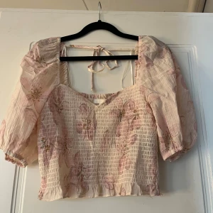 Blommig blus - Söt blommig blus i ljusrosa med puffiga ärmar och knytband i ryggen. Blusen har en smockad design och volangdetaljer vid fållen. Perfekt för en romantisk look.