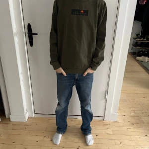 Olivgrön sweatshirt från Ellesse - Säljer en snygg olivgrön sweatshirt från Ellesse i mycket bra skick. Den har en klassisk rund hals och långa ärmar, perfekt för både höst och vår. Loggan är tryckt på bröstet i svart och orange. Superbekväm och stilren, passar till allt! 🌿