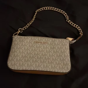 Beige axelväska från Michael Kors - Snygg beige axelväska från Michael Kors med kedjeaxelrem och dragkedja. Väskan har ett diskret mönster och är perfekt för att bära dina nödvändigheter. Insidan är rymlig med flera fack för enkel organisering.