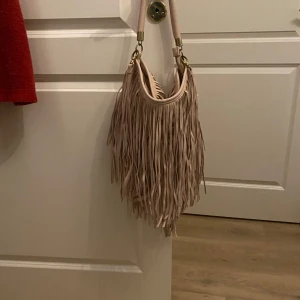 Beige axelväska med fransar - Säljer en snygg beige axelväska med långa fransar. Väskan har en justerbar axelrem och är perfekt för att ge din outfit en bohemisk touch. Den är rymlig och passar för alla dina vardagsbehov.
