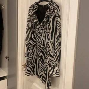 Zebramönstrad klänning från Boohoo - Snygg zebramönstrad klänning från Boohoo med volangdetaljer och dragkedja i ryggen. Klänningen har långa ärmar och en v-ringad design som ger en trendig look. Perfekt för att sticka ut med stil.