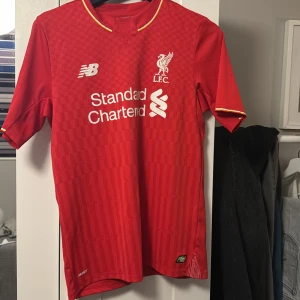 Röd Liverpool fotbollströja från New Balance - Säljer en röd Liverpool fotbollströja med korta ärmar. Tröjan har klubbens emblem och sponsorlogga på framsidan samt spelarnamnet Benteke och nummer 9 på baksidan. En riktigt snygg retro tröja! Köpt för många år sedan men har använt sparsamt. 