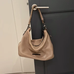 Beige axelväska från Christina Christensen - Snygg beige axelväska i skinn från Christina Christensen. Väskan har en stilren design med en praktisk axelrem och en diskret logotyp på framsidan. Perfekt för att bära med sig det nödvändigaste med stil. Äkta kalvskinn 