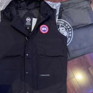 Svart dunväst från Canada Goose - Säljer en svart dunväst från Canada Goose med det ikoniska märket på bröstet. Västen har både dragkedja och knappar framtill för extra skydd mot kyla. Perfekt för om man vill hålla sig varm men samtidigt stilig