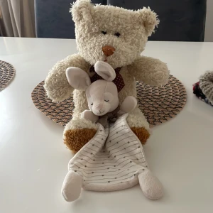 Gosedjur och snuttefilt - Säljer ett mjukt och gosigt gosedjur i form av en nalle med brun rosett och bruna tassar. Medföljer en snuttefilt med kaninhuvud, vit med grå ränder och små stjärnor. Perfekt för barn som behöver en trygg kompis.