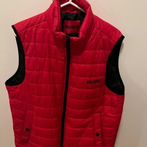 Hugo boss väst st-M - Snygg röd quiltad väst från Hugo med svarta detaljer. Västen har en dragkedja framtill och två fickor med knappar. Perfekt för att hålla värmen under kyliga dagar.
