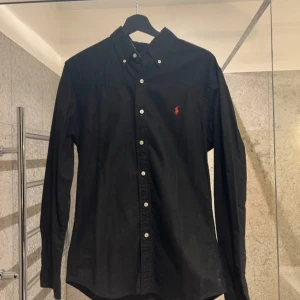 Svart skjorta från Ralph Lauren - Snygg svart skjorta från Ralph Lauren med klassisk krage och knappar framtill. Den har en broderad logotyp i rött på bröstet och långa ärmar. Perfekt för en stilren look.