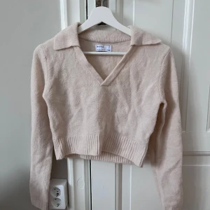 Beige croppad tröja från Bershka - Säljer en beige croppad tröja från Bershka med krage och v-ringning. Tröjan har långa ärmar och ribbade detaljer vid nederkanten och ärmsluten. Perfekt för en stilren look!