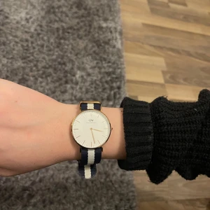 Randig klocka från Daniel Wellington - Snygg klocka från Daniel Wellington med randigt armband i blått och vitt. Klockan har en stilren urtavla och guldfärgade detaljer. Perfekt accessoar för en klassisk look. Den är nyskick och knappt använd då jag inte bär klocka. Nypris 1799