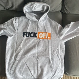 Grå hoodie med tryck - Säljer en grå hoodie med texten 'FUCK OFF' i svart och orange på framsidan. Tröjan har en klassisk känguruficka och en bekväm huva. Perfekt för en avslappnad stil.