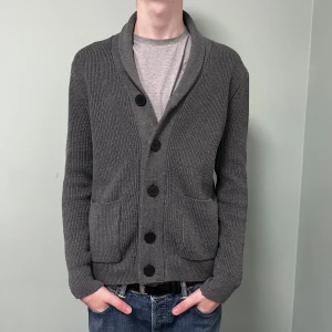 Cardigan med knappar - Säljer en stilren grå cardigan med stora svarta knappar och två praktiska fickor framtill. Modellen är 195 och Storlek L. Hör av er vid intresse!