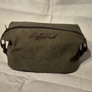 Olivgrön necessär med dragkedja - Snygg olivgrön necessär med broderad text på framsidan. Praktisk dragkedja upptill och robust design, perfekt för att hålla ordning på dina saker. Den har även mycket bra fack inuti så du håller ordning på dina saker. 