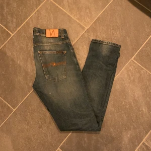 Blå jeans från Nudie Jeans - Snygga blå jeans från Nudie Jeans med klassisk femficksdesign och orange sömmar. Jeansen har en normal passform och är tillverkade i slitstarkt denim. Perfekta för en avslappnad stil. 