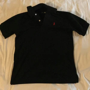 Svart pikétröja från Ralph Lauren - Snygg svart pikétröja från Ralph Lauren med klassisk krage och knappslå. Den har en röd broderad logotyp på bröstet som ger en stilren touch. Perfekt för en avslappnad men ändå stilfull look.