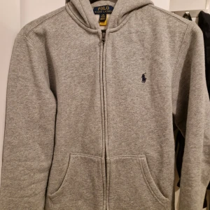 Grå hoodie från Ralph Lauren - Säljer en stilren grå hoodie från Ralph Lauren med dragkedja och fickor framtill. Den har en klassisk design med det ikoniska logotypen broderad på bröstet. Perfekt för en avslappnad look.