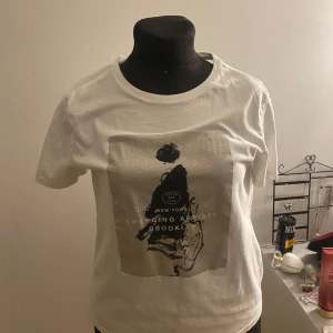 Vit t-shirt med tryck - Snygg vit t-shirt med ett stilrent tryck av en person och texten 'New York Emerging Artists Brooklyn'. Perfekt för en avslappnad look.