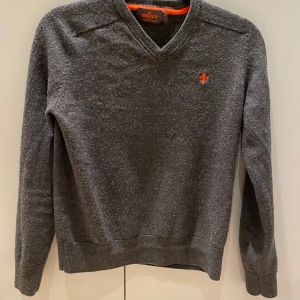 Grå tröja från Morris - Snygg grå tröja från Morris med v-ringning och en liten orange broderad detalj på bröstet. Perfekt för en stilren look. Tröjan har långa ärmar och ribbade muddar vid ärmslut och nederkant. Storlek 140 - 150