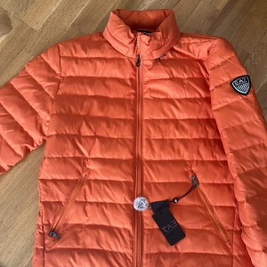 Orange quiltad jacka från EA7 - Snygg orange quiltad jacka från EA7 med dragkedja och hög krage. Perfekt för kyligare dagar med sin pufferdesign och stilrena look. Jackan har två praktiska fickor med dragkedja och en logotyp på ärmen.